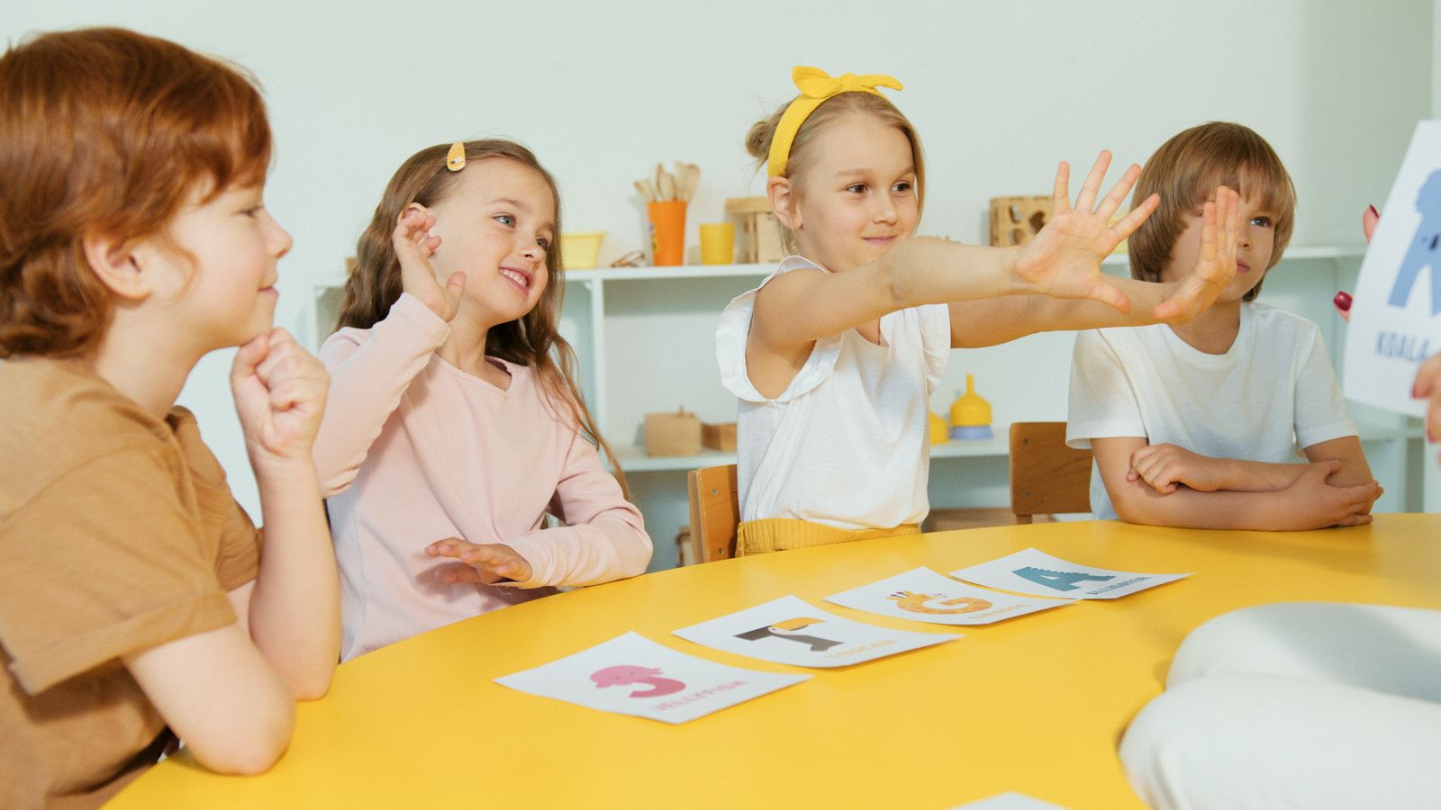 Children around a yellow table eagerly raising their hands during a picture-card game