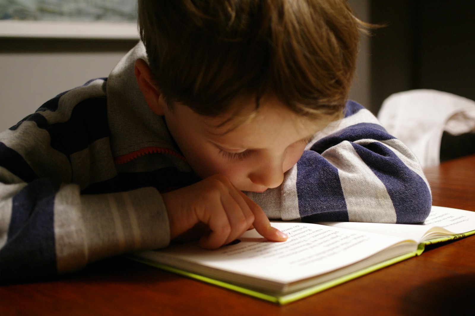 A child leaning over a table, following the lines of a book with a finger